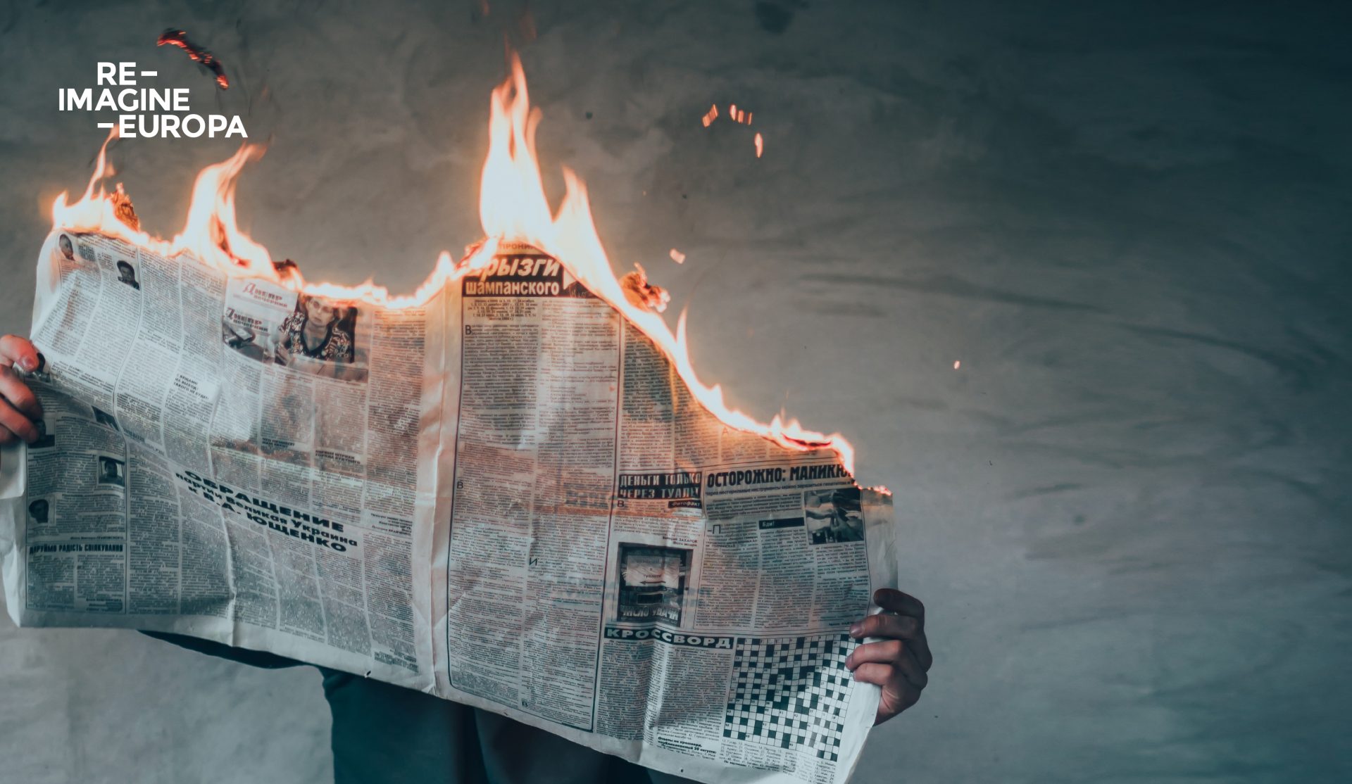 Image of a man holding a newspaper on fire with a grey wall on the background