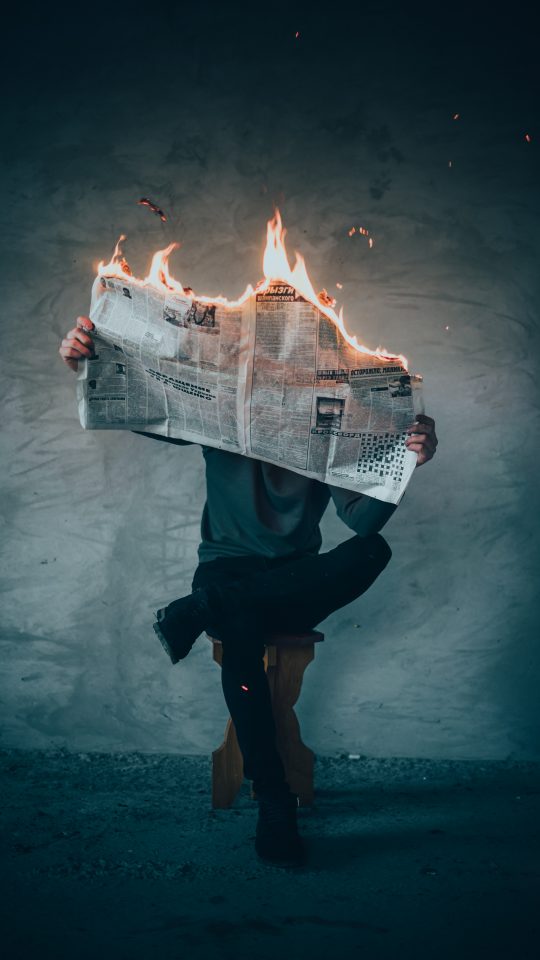Man seated in a wooden bench holding an open newspaper on fire